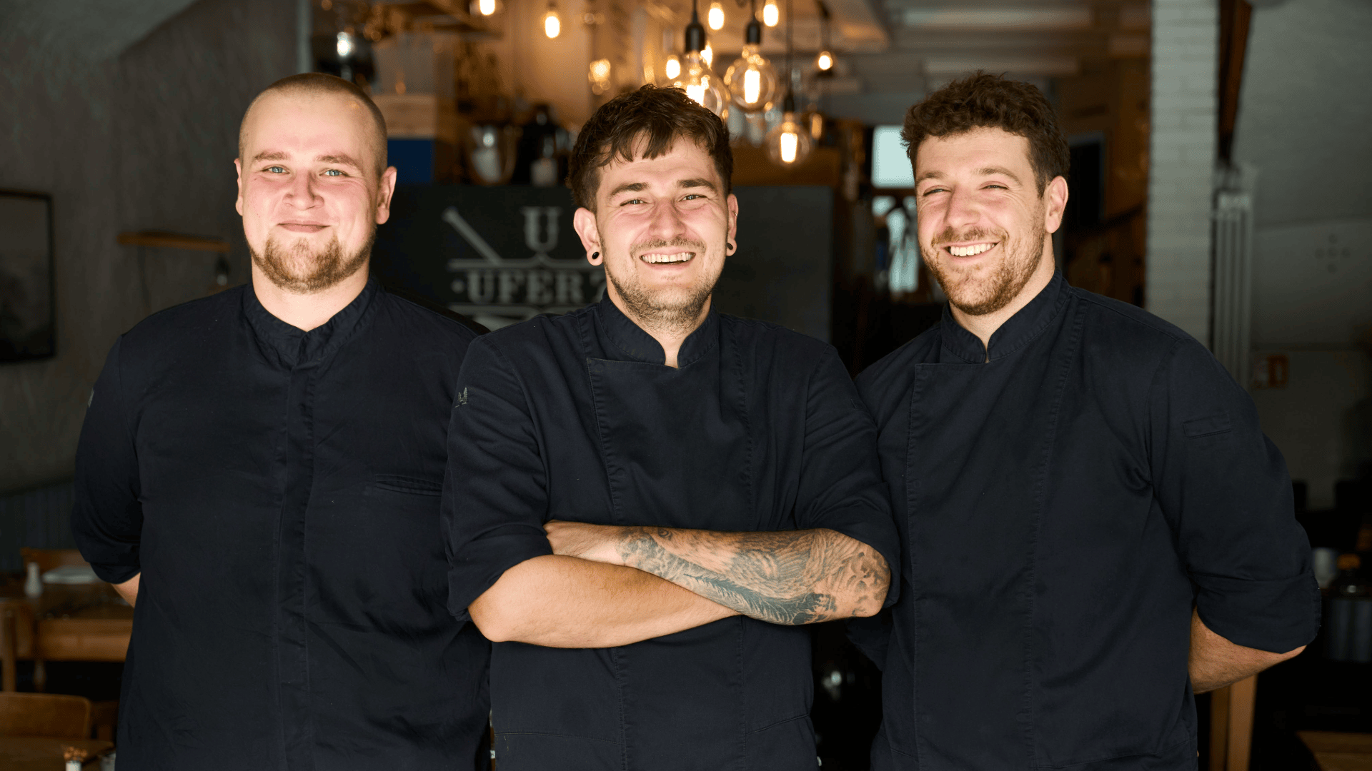 Drei lächelnde Köche in dunklen Kochuniformen stehen gemeinsam in einem Restaurant.