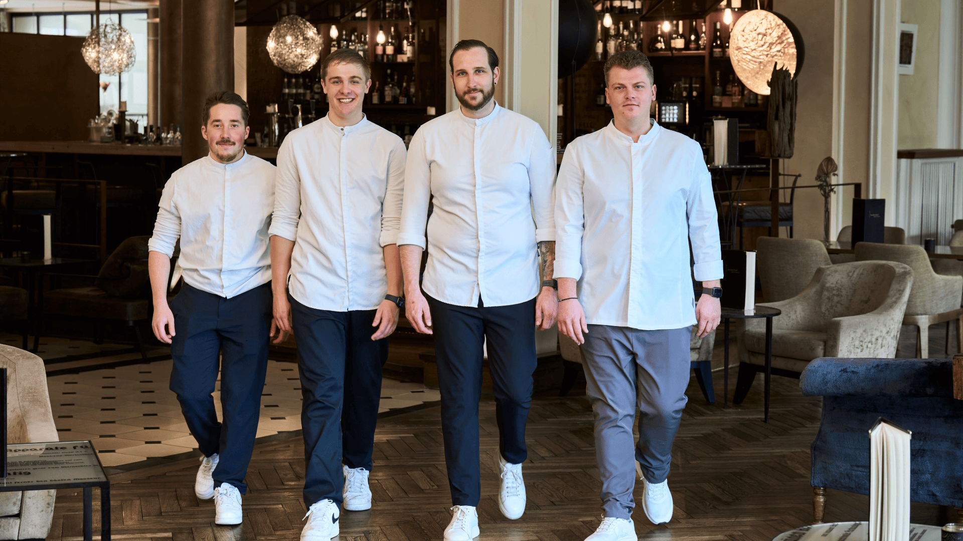 Vier Männer in weißen Langarmhemden und Freizeithosen gehen in einer Reihe durch ein Restaurant.
