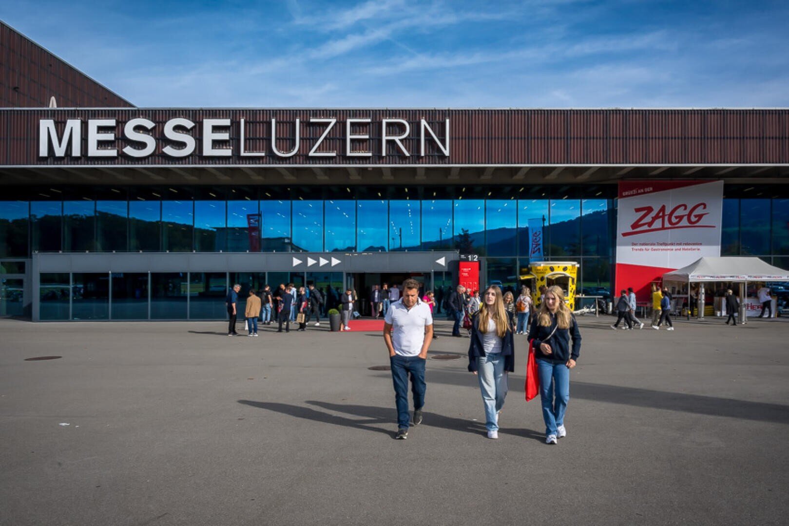 Vor dem Messezentrum „Messe Luzern“ gehen Menschen vorbei, über ihnen hängt ein großes Schild, während andere das Gebäude betreten. Im Hintergrund sind ein rot-weißes ZAGG-Banner und ein gelber Bus zu sehen.