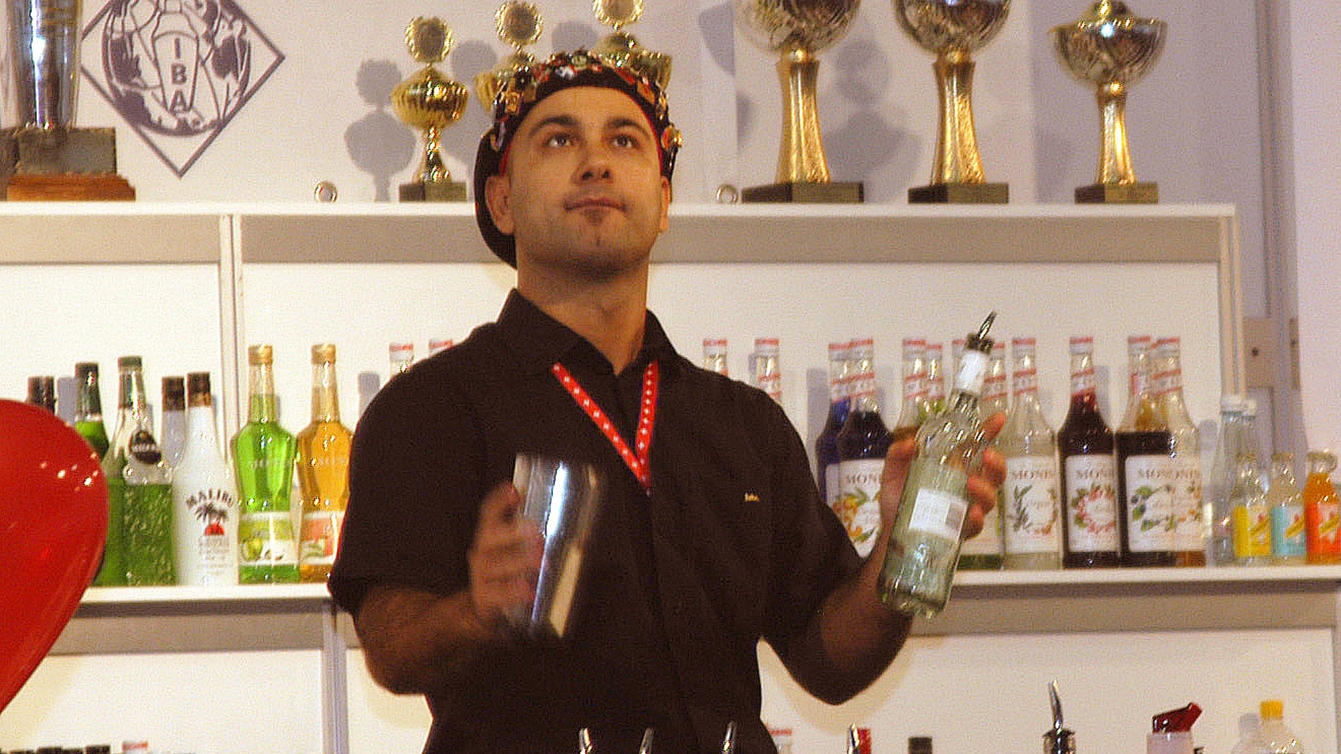 Ein Barkeeper in einem schwarzen Hemd und mit einem Blumenkranz im Haar hält einen Cocktailshaker und eine Spirituosenflasche in der Hand; hinter ihm stehen auf Regalen Pokale und verschiedene Flaschen.