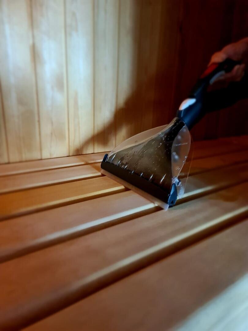 Eine Hand reinigt mit einem Teppichreiniger die Holzbank in einer Sauna; im durchsichtigen Saugkopf des Geräts ist Wasser zu sehen.