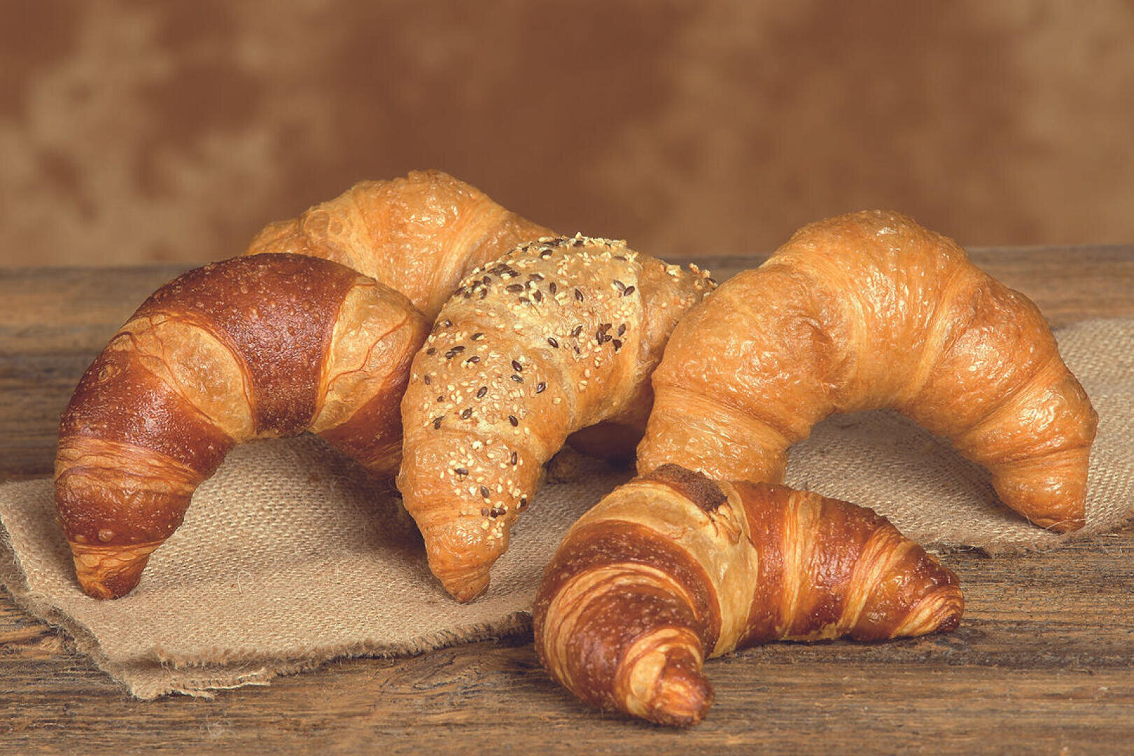Fünf Croissants, darunter sowohl einfache als auch mit Körnern bestreute Varianten, sind auf einem Stück Sackleinen auf einer rustikalen Holzunterlage angeordnet, vor einem braunen, unscharfen Hintergrund.