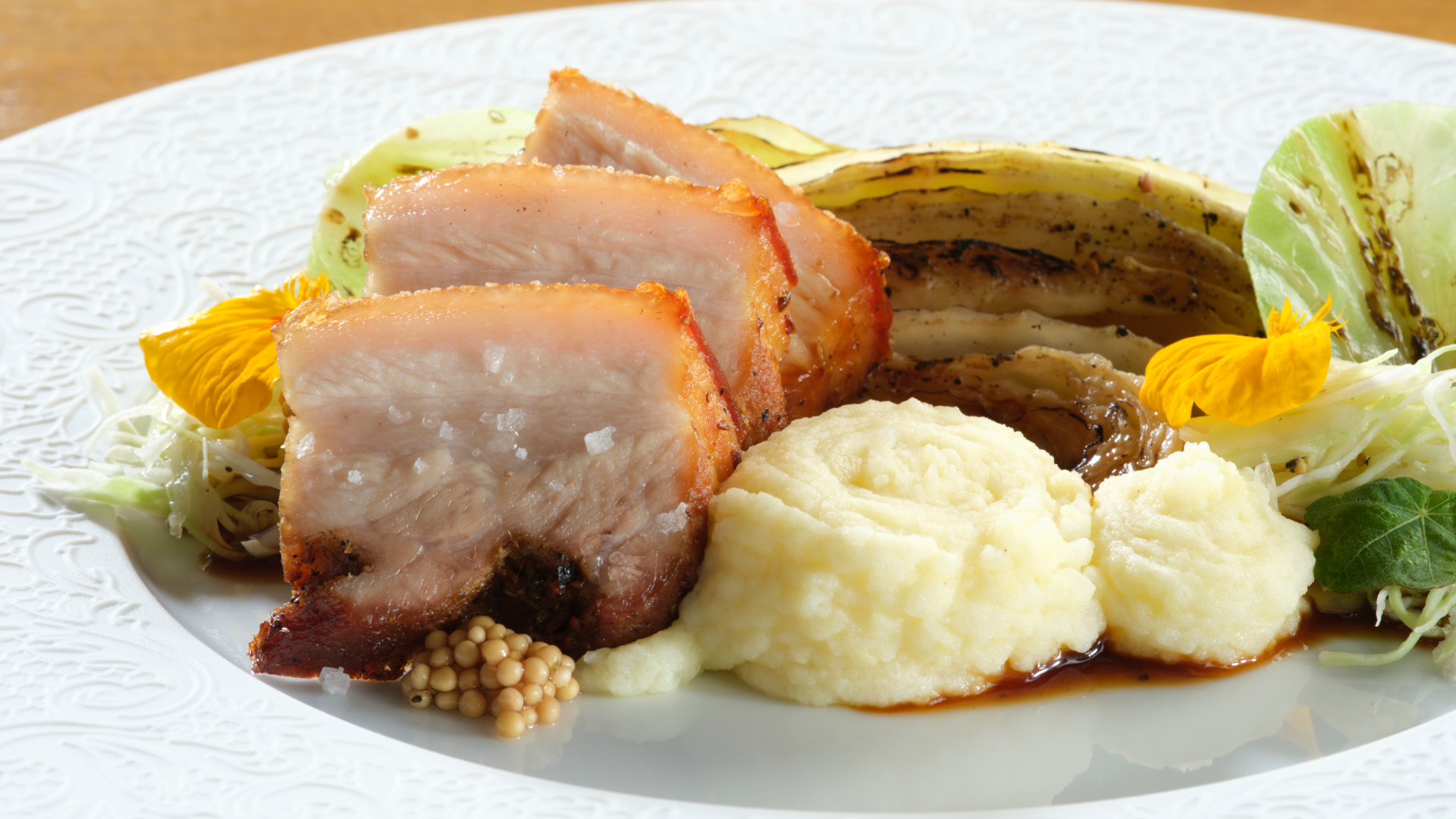 Scheiben von gebratenem Schweinebauch mit Kartoffelpüree, angebratenem Kohl und Senfkörnern auf einem weißen Teller.
