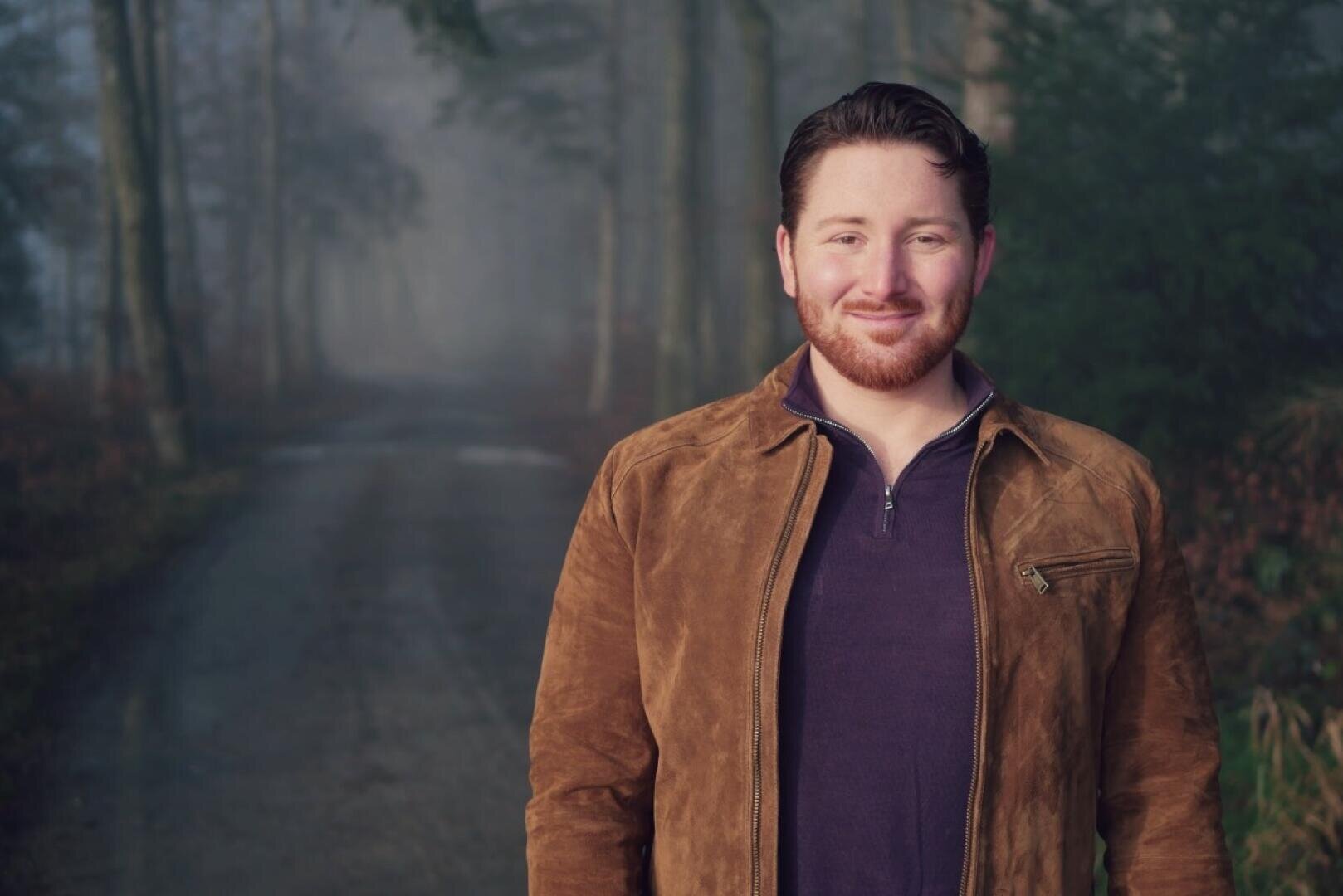 Ein Mann mit rotem Bart, der eine braune Jacke über einem dunkelvioletten Hemd trägt und auf einer nebligen Straße steht.
