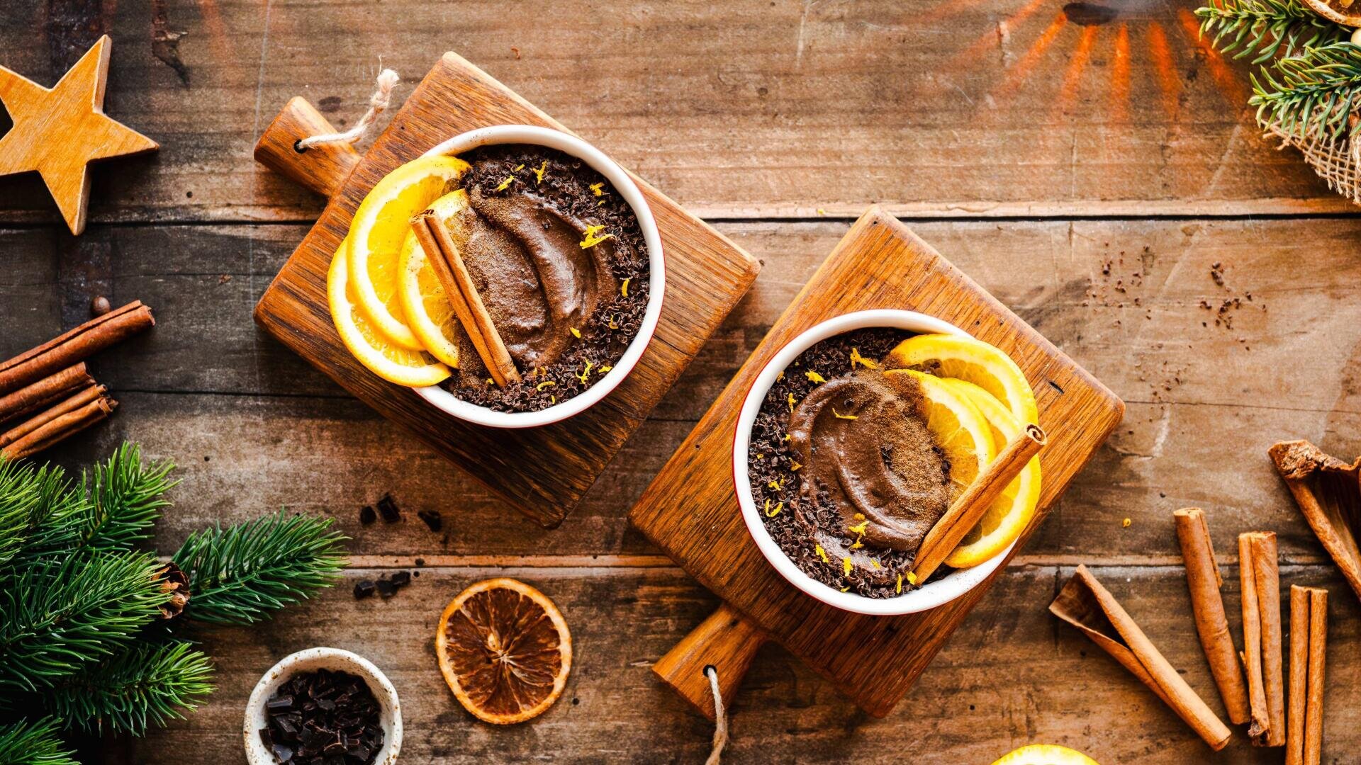 Zwei Schalen mit Mousse aus dunkler Schokolade, serviert auf Holzschneidebrettern. Jede ist mit frischen Orangenscheiben, einer Zimtstange und Schokoladenraspeln garniert. Die Szene spielt sich auf einem rustikalen Holztisch ab, der mit Kiefernzweigen, Zimt und getrockneten Orangen verziert ist.