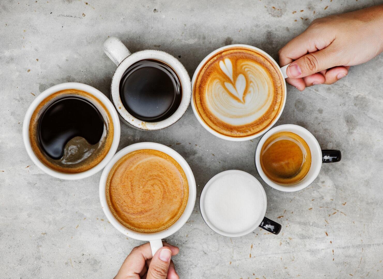 Aufnahme von oben auf sechs verschiedene Kaffeegetränke in Tassen auf einer grauen Fläche, wobei Hände zwei der Tassen halten.