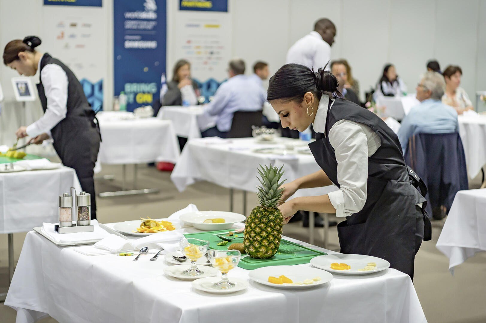 Eine Frau in einer schwarzen Schürze schneidet an einem Tisch in einer hellen, professionellen Umgebung eine Ananas in Scheiben. Andere Menschen arbeiten oder sitzen an benachbarten Tischen, und auf dem Tisch stehen Teller mit Obst.