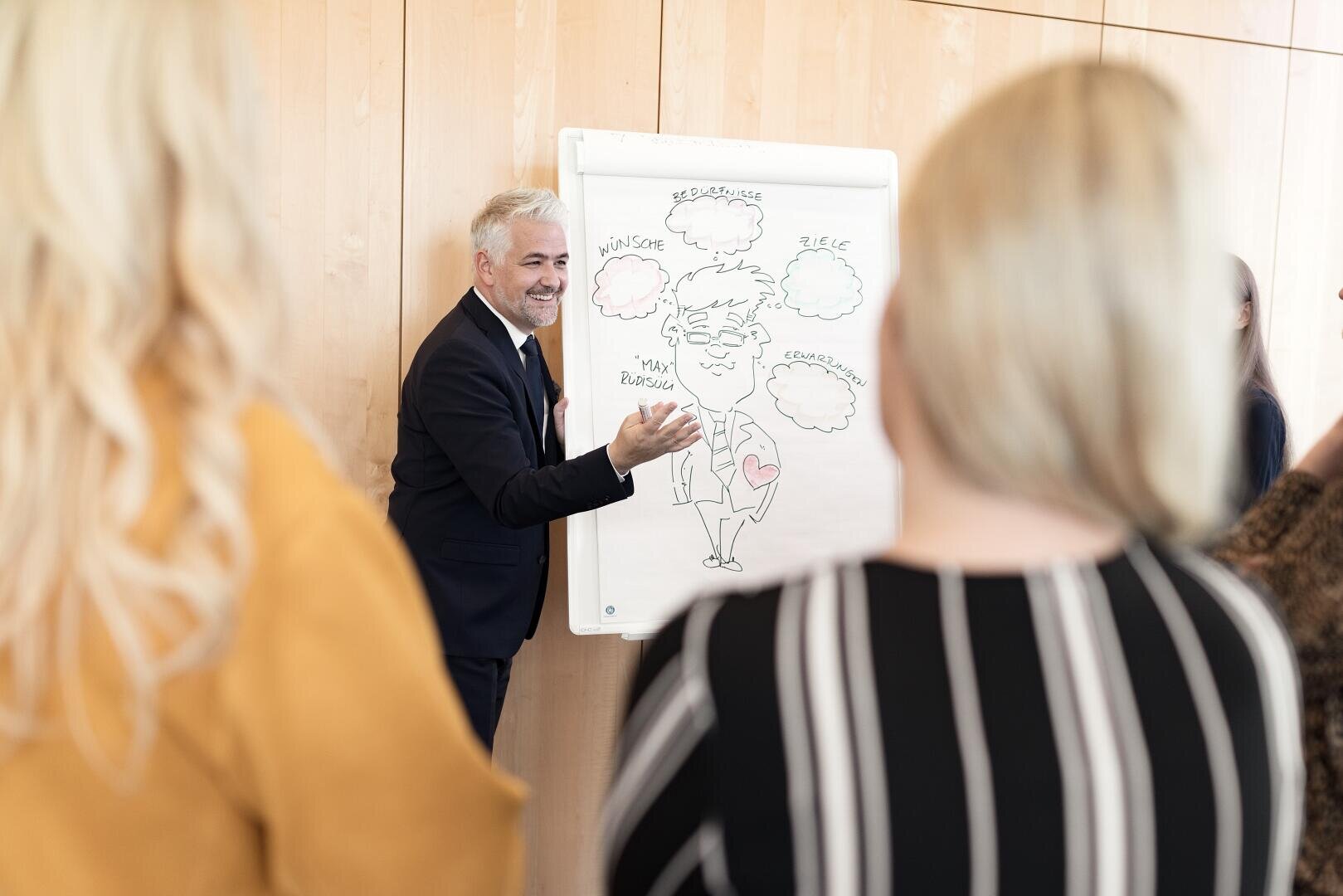 Ein Mann im Anzug steht neben einem Flipchart, auf dem eine Karikatur und Sprechblasen zu sehen sind. Er lächelt und unterhält sich mit drei Personen, die der Kamera den Rücken zuwenden.
