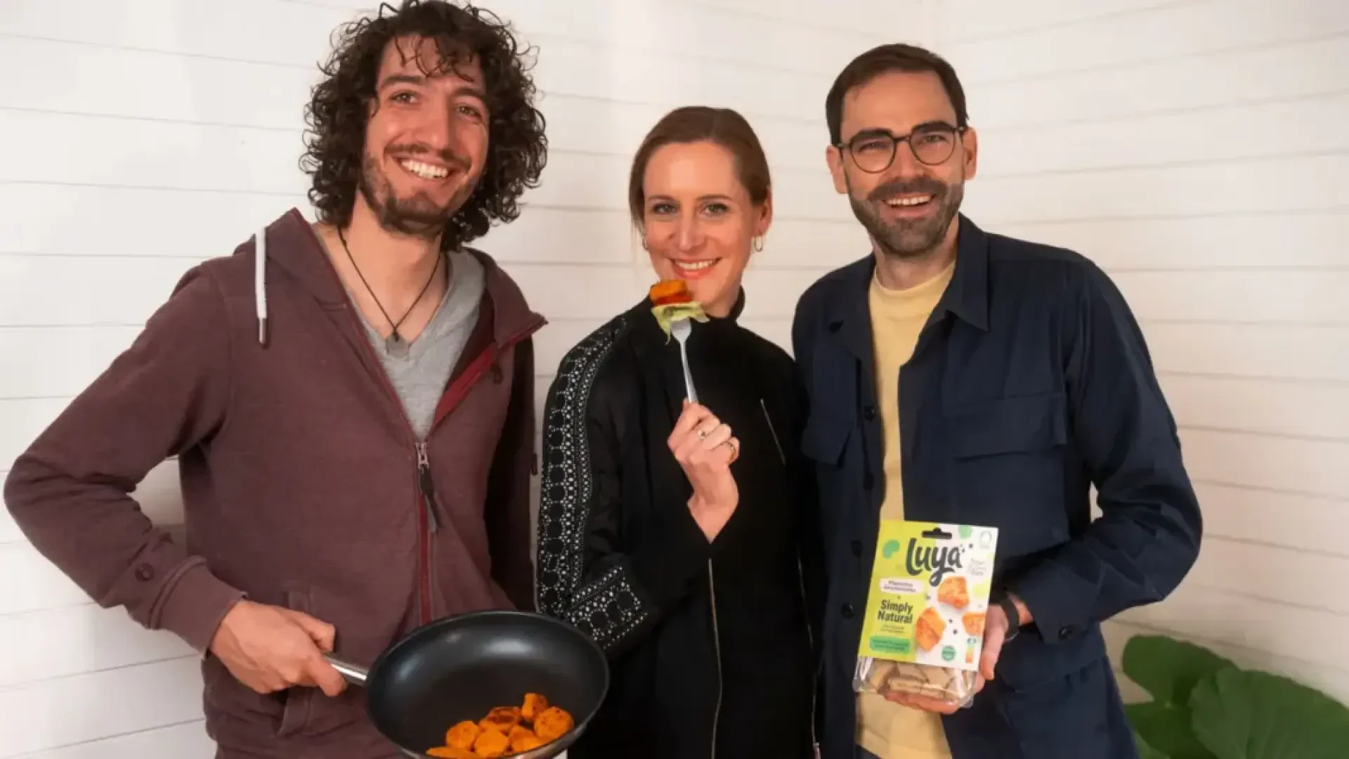 Drei Personen lächeln, während sie eine Bratpfanne mit fertig zubereitetem Essen, eine Gabel mit einem Stück Essen und eine Packung Luya Simply Natural in den Händen halten.