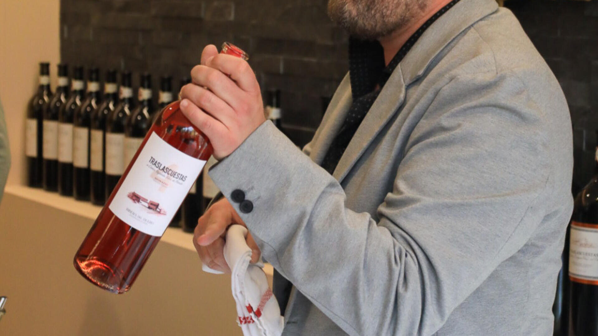 Eine Person in einem grauen Blazer hält eine Flasche Roséwein und ein weißes Handtuch in der Hand. Im Hintergrund sind auf einem Regal vor einer dunklen Wand mehrere Flaschen Wein zu sehen.