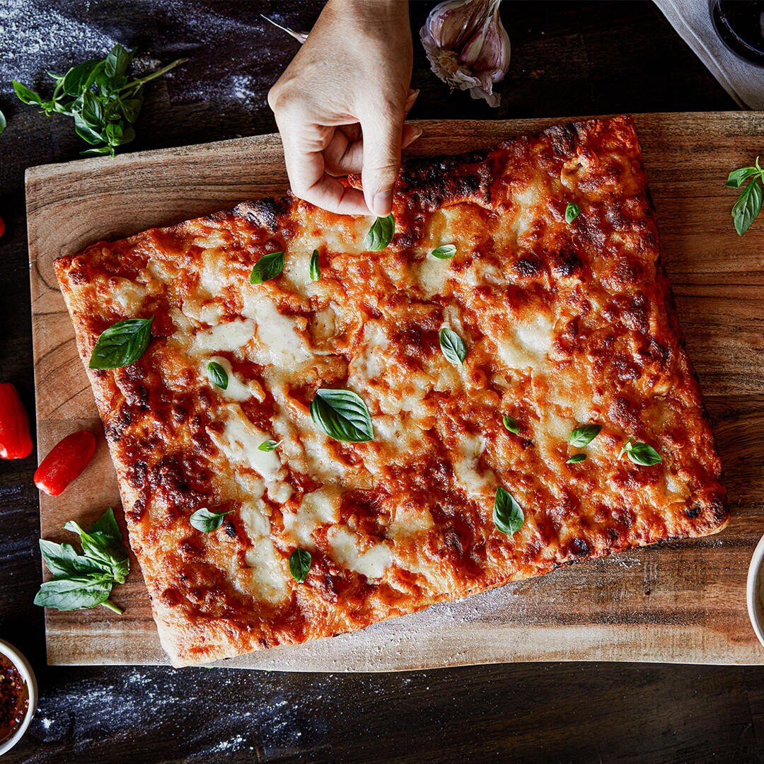 Eine Hand streut frische Basilikumblätter auf eine rechteckige Käsepizza auf einem Holzbrett, umgeben von Knoblauch, Kräutern und Chiliflocken.