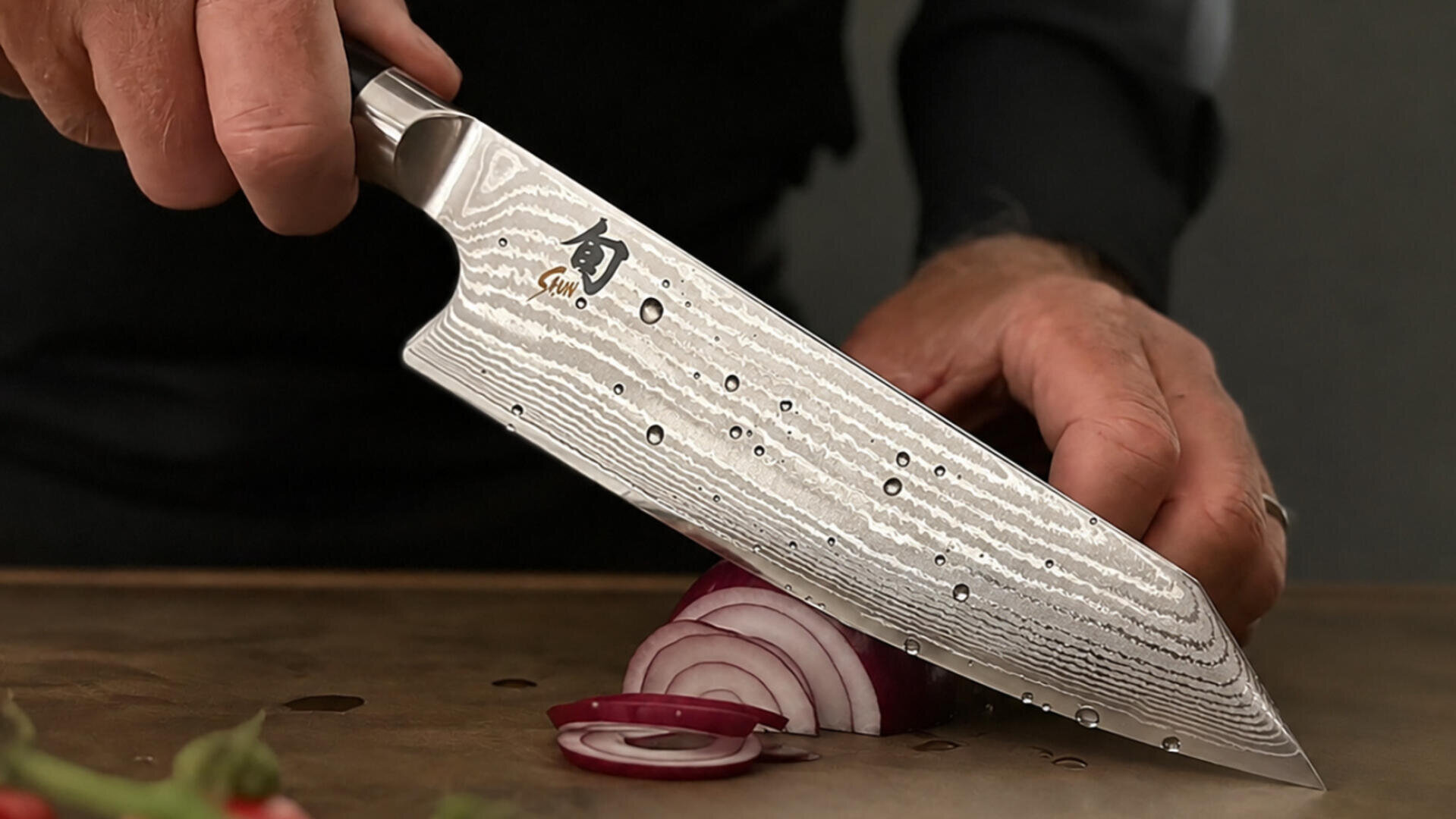 Eine Person schneidet mit einem großen, gemusterten Kochmesser auf einem Schneidebrett eine rote Zwiebel in Scheiben. Das Messer hat ein markantes, wellenförmiges Muster auf der Klinge.