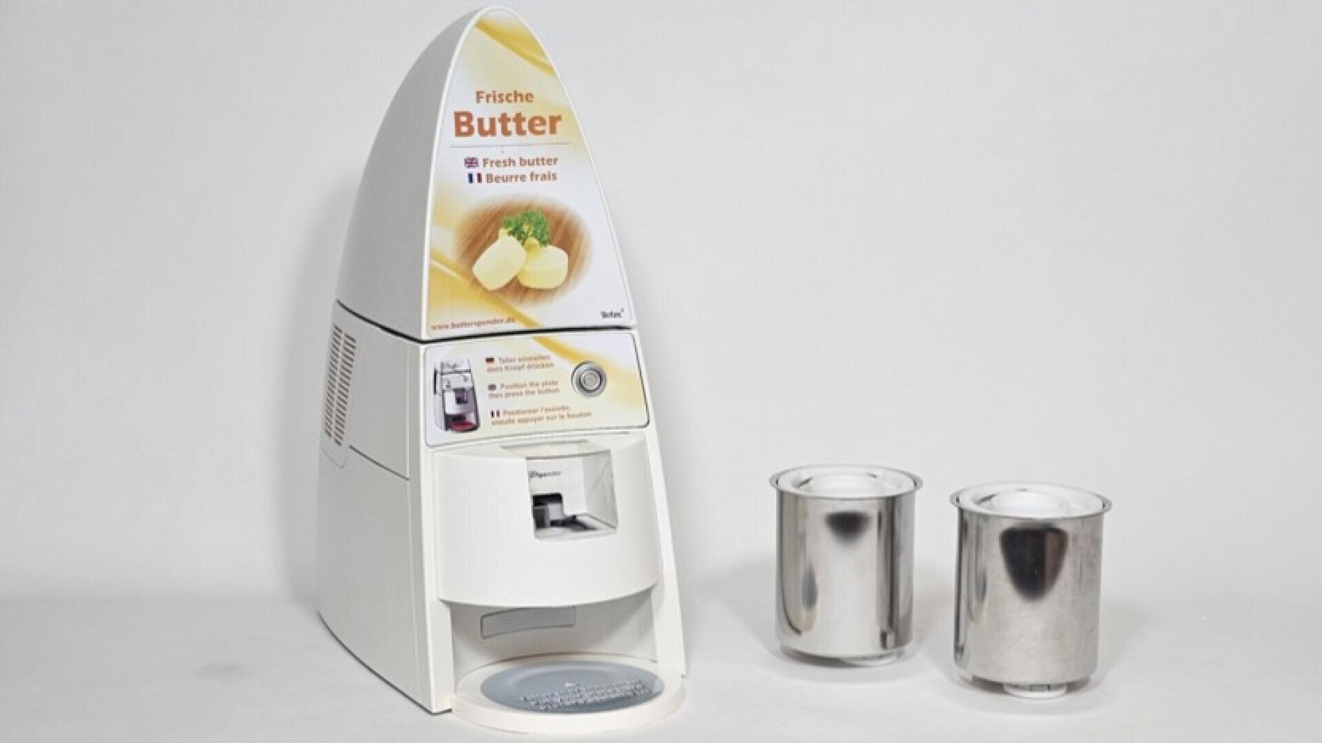 Auf einem schlichten weißen Hintergrund steht ein weißer Butterbehälter mit einer Abbildung, die Butter auf einem Teller zeigt, neben zwei Metallbehältern.
