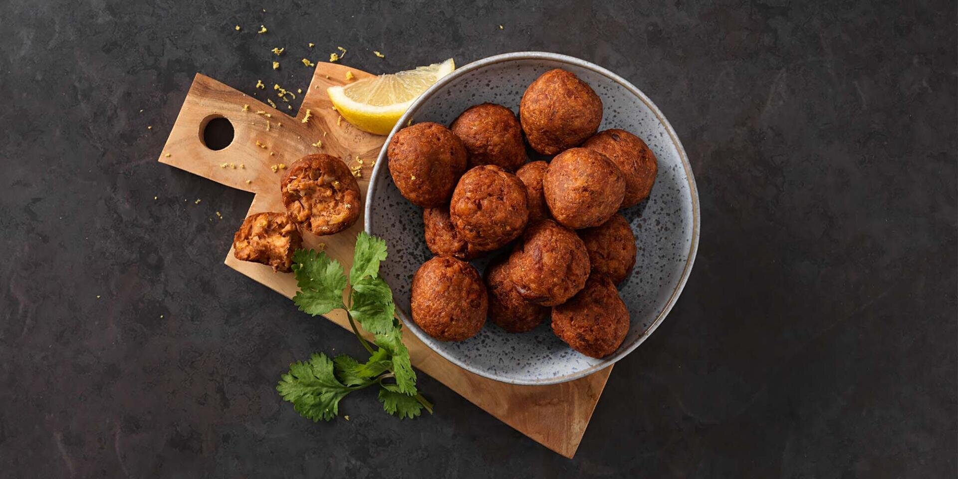 Eine Schale mit runden, goldbraunen Falafelbällchen auf einem Holzbrett, dazu eine Zitronenscheibe, Koriander und ein zerbrochenes Falafelbällchen – alles auf einer dunklen, strukturierten Oberfläche.