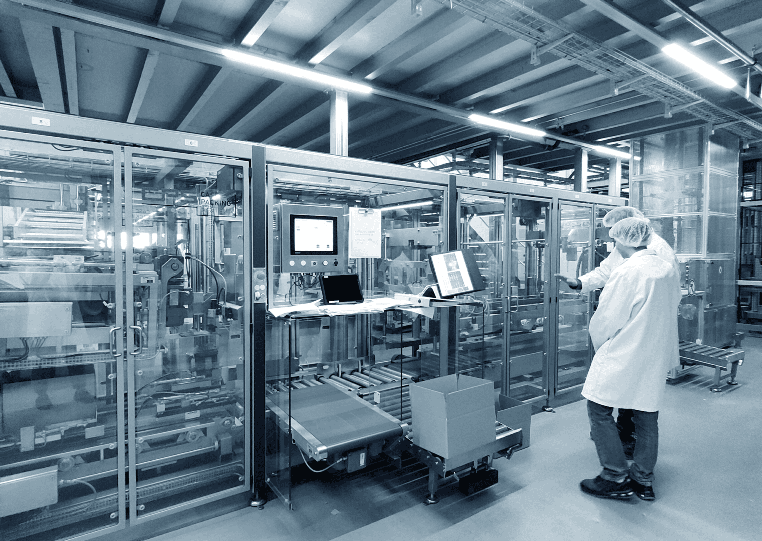 Zwei Personen in Laborkitteln und Haarnetzen stehen vor automatisierten Maschinen mit Computern und Kontrollbildschirmen in einer industriellen Fabrik oder einem Labor.