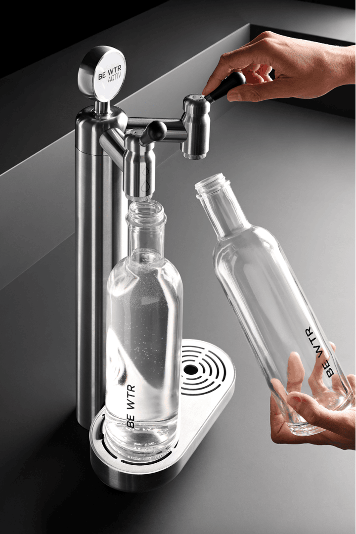 Eine Person füllt eine Glasflasche mit Wasser aus einem modernen Wasserspender aus Edelstahl mit der Aufschrift „BE WTR“. Eine weitere leere Flasche steht daneben auf einer eleganten schwarzen Arbeitsplatte.