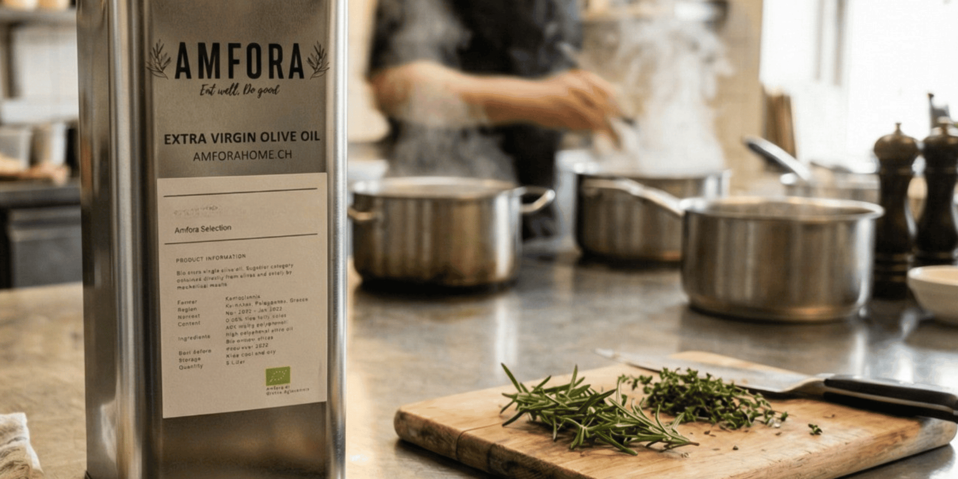 Eine Metalldose mit Amfora-Olivenöl extra vergine steht auf einer Küchenarbeitsplatte neben einem Holzschneidebrett mit frischen Kräutern und einem Messer, während im Hintergrund ein Koch am Herd steht.