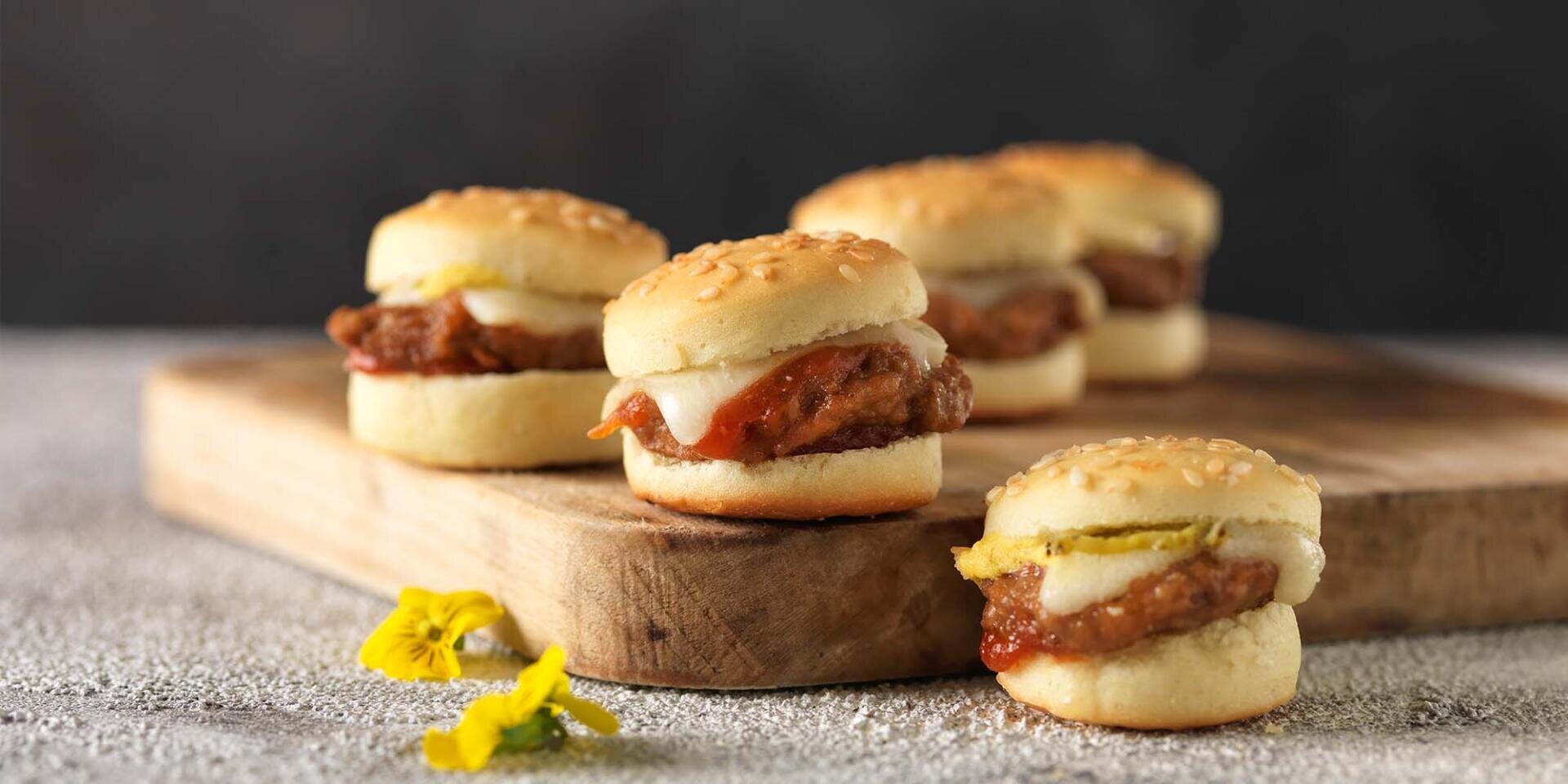 Auf einem Holzbrett sind vier Mini-Burger mit Sesambrötchen, Fleischpasteten, Käse und Beilagen angerichtet. Im Vordergrund liegen neben den Burgern zwei gelbe essbare Blüten.