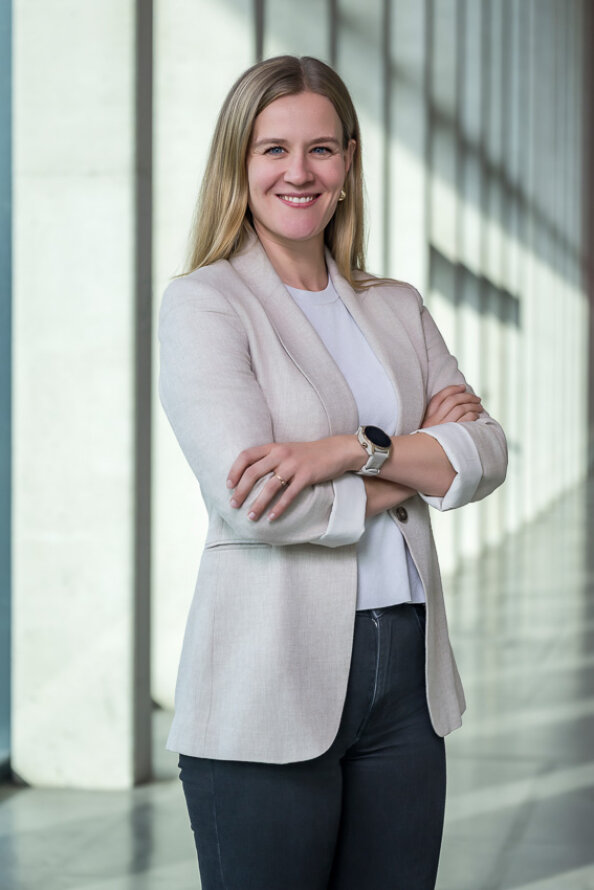 Eine Frau mit langen blonden Haaren steht mit verschränkten Armen in einem Haus und lächelt. Sie trägt einen hellen Blazer, ein weißes Oberteil und eine dunkle Hose. Im Hintergrund sind hohe Fenster und geometrische Schatten zu sehen.