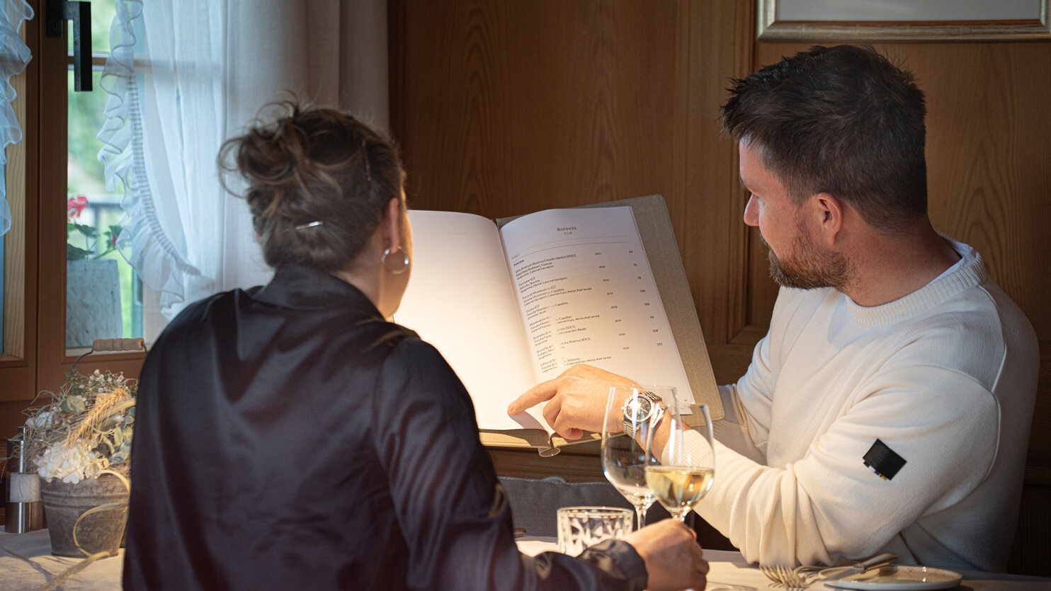 Ein Mann und eine Frau sitzen an einem Tisch in einem gemütlichen Restaurant und schauen sich gemeinsam die Speisekarte an. Der Mann zeigt auf die Speisekarte, während die Frau zuhört. Auf dem Tisch stehen Weingläser und ein Blumenstrauß.
