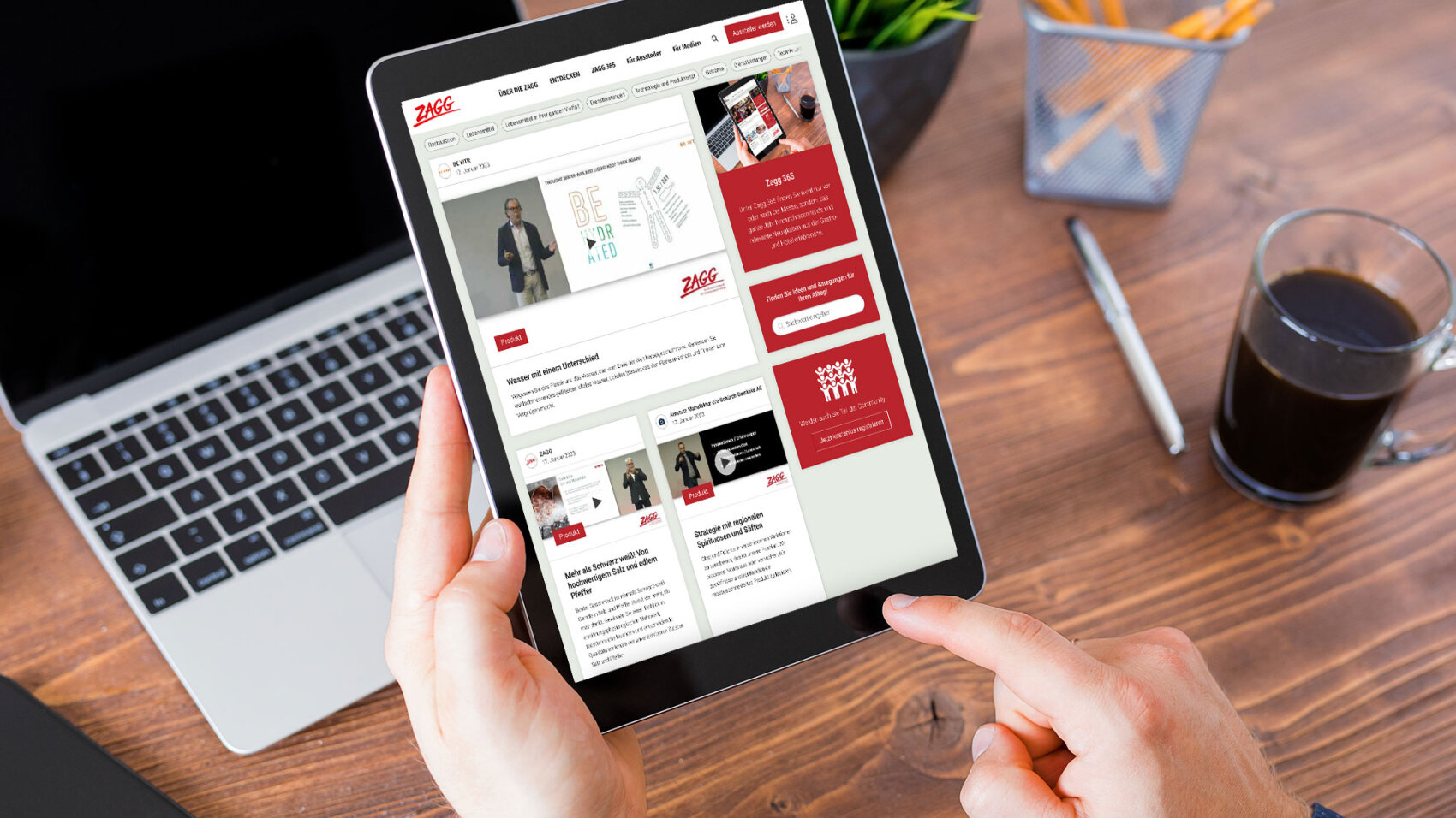 Eine Person hält ein Tablet in der Hand, auf dem eine professionelle Website mit Artikeln und Bildern zu sehen ist. Auf dem Holztisch im Hintergrund sind ein Laptop, eine Tasse Kaffee und ein Stifthalter zu sehen.