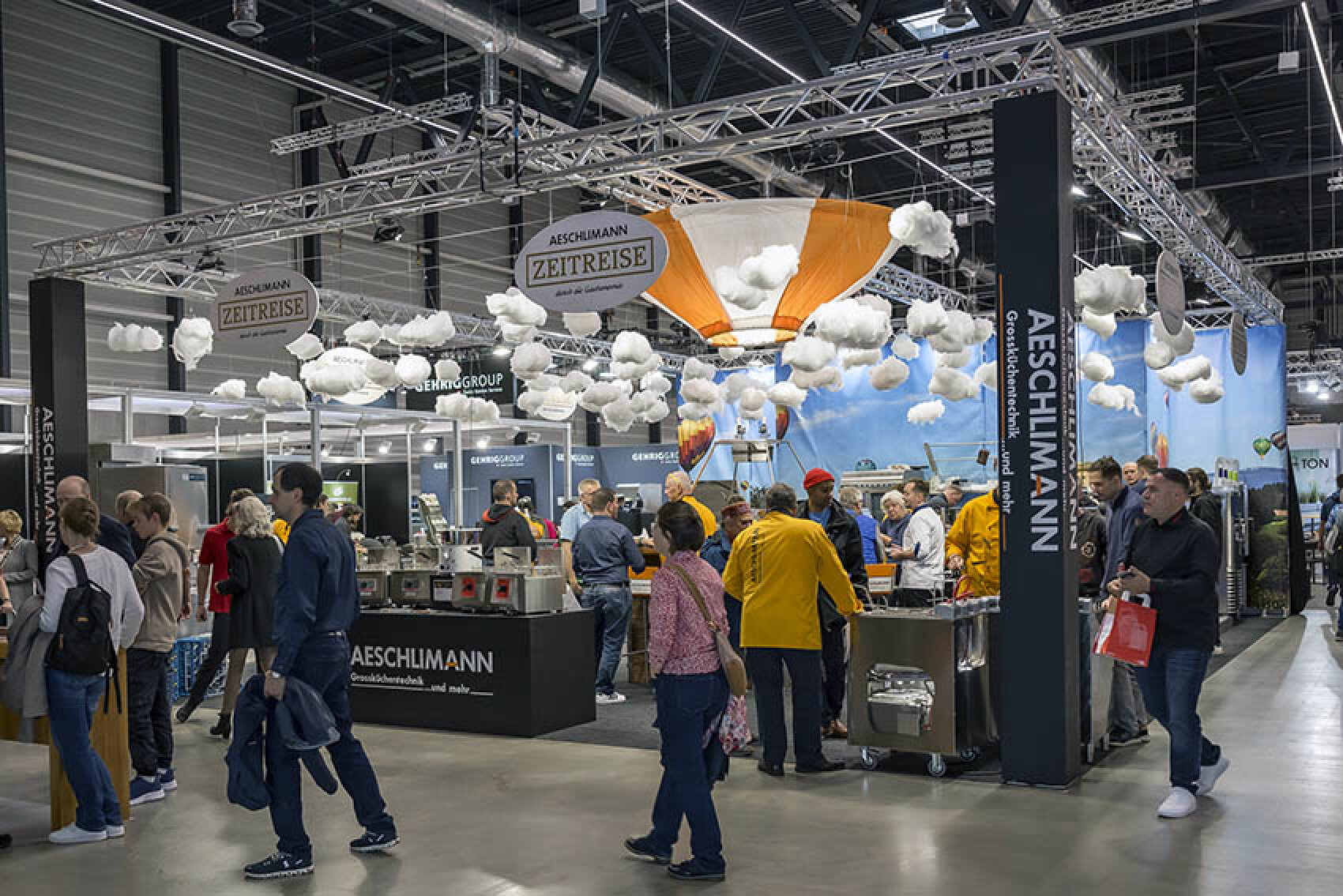Ein belebter Messestand mit Menschen, die sich um Auslagen und Theken scharen. Der Stand ist mit hängenden Wolkendekorationen und einem großen Heißluftballon dekoriert, auf dessen Bannern "Aeschlimann Zeitreise" steht.